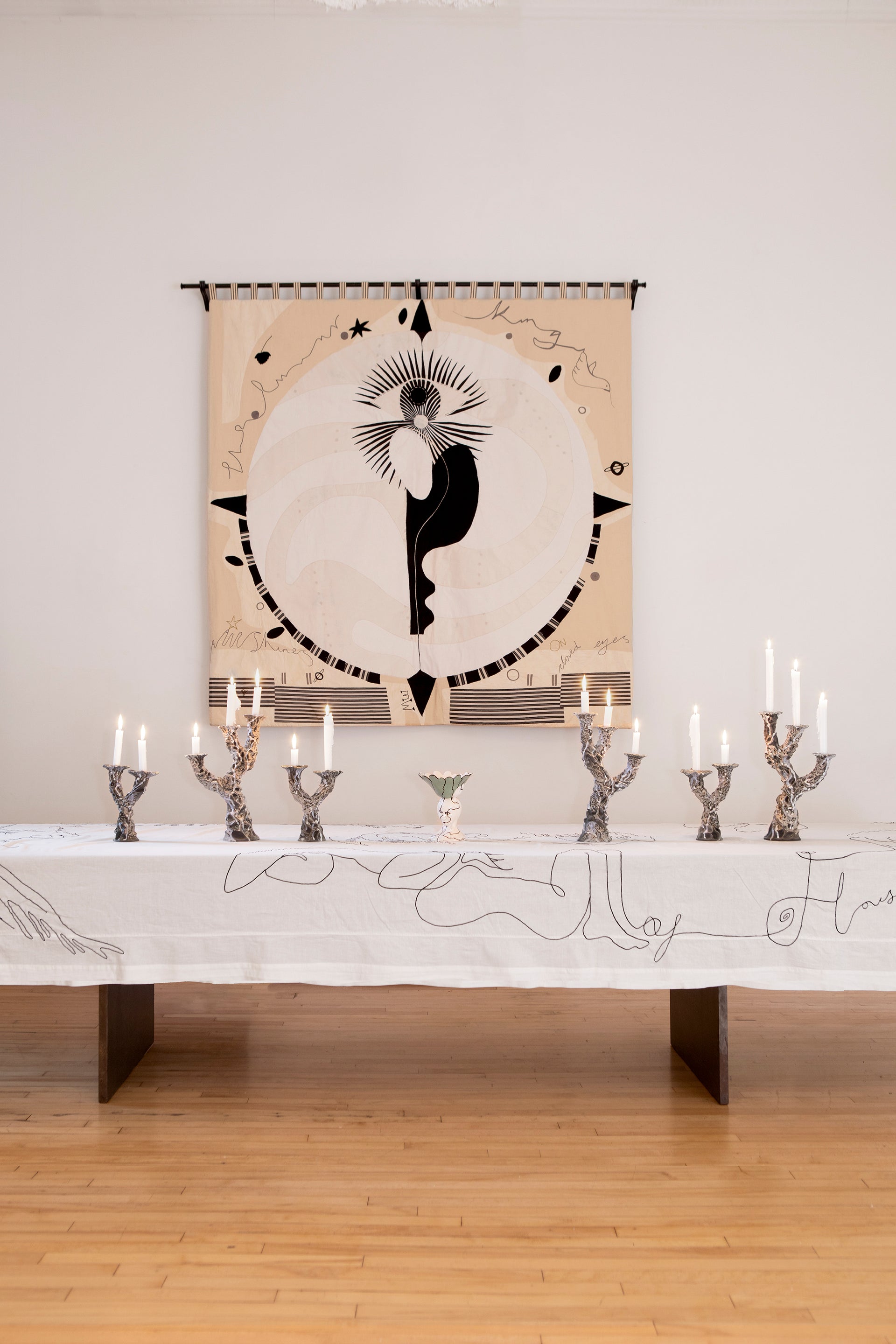 Hand made large decorative tapestry with an abstract and surrealist design motif. A moon design with radiating stars and celestial patterns and line work in an ivory and cream palette. Hand crafted and hand embroidered from antique silks. Made in India, artisanal home decor. Modern tapestry design. Shown in a home setting with modern ceramic objects on a table setting. 