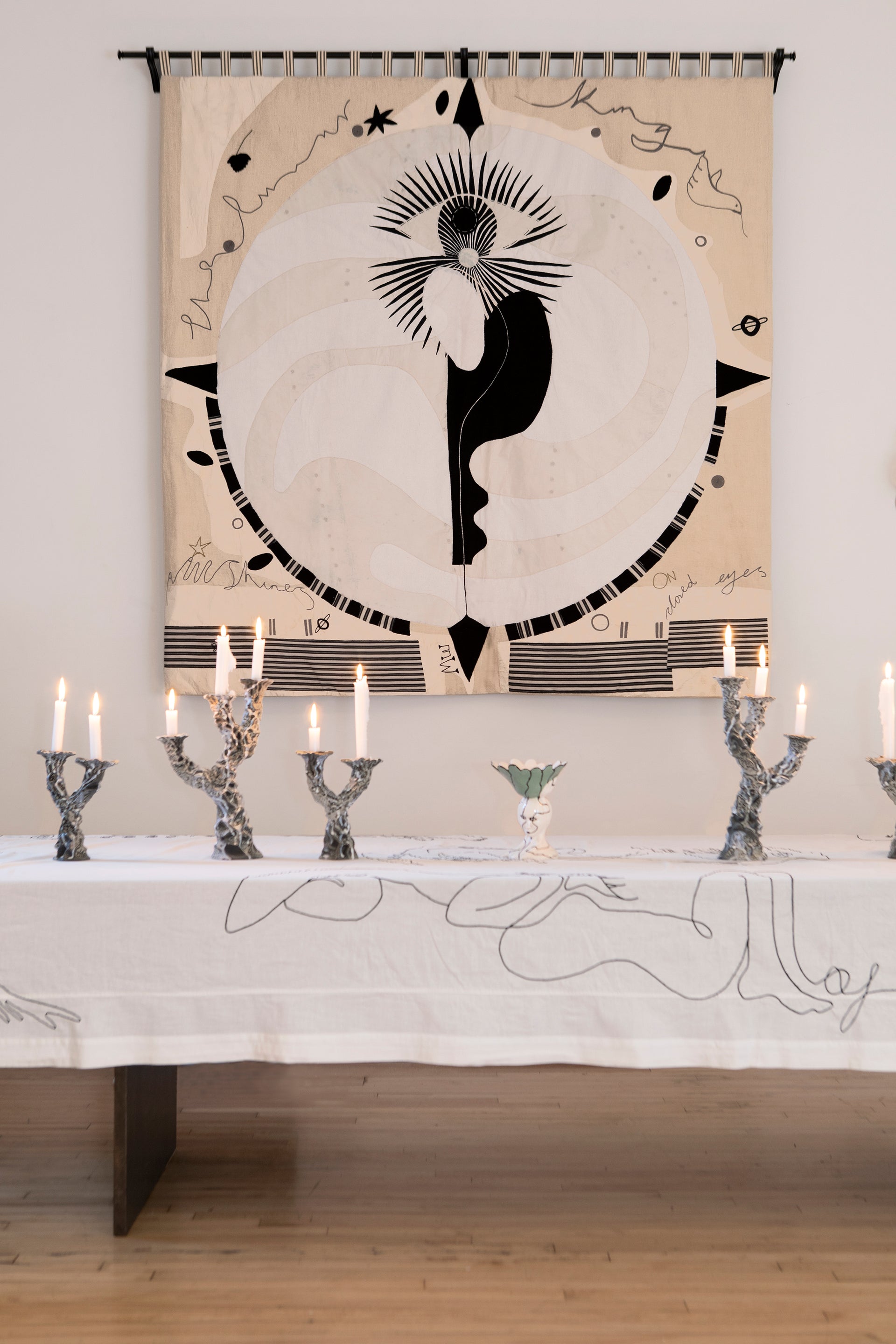 Hand made large decorative tapestry with an abstract and surrealist design motif. A moon design with radiating stars and celestial patterns and line work in an ivory and cream palette. Hand crafted and hand embroidered from antique silks. Made in India, artisanal home decor. Modern tapestry design. Shown in a home setting with modern ceramic objects on a table setting. 