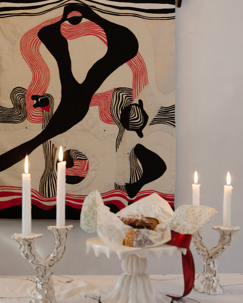 Hand made decorative tapestry with an abstract and surrealist design motif. Swirling abstract line work in bold red and black on an ivory background. Hand crafted and hand embroidered from antique silks. Made in India, artisanal home decor. Modern tapestry design. Shown in a home setting with a set table of ceramic objects, set for a dinner party. 
