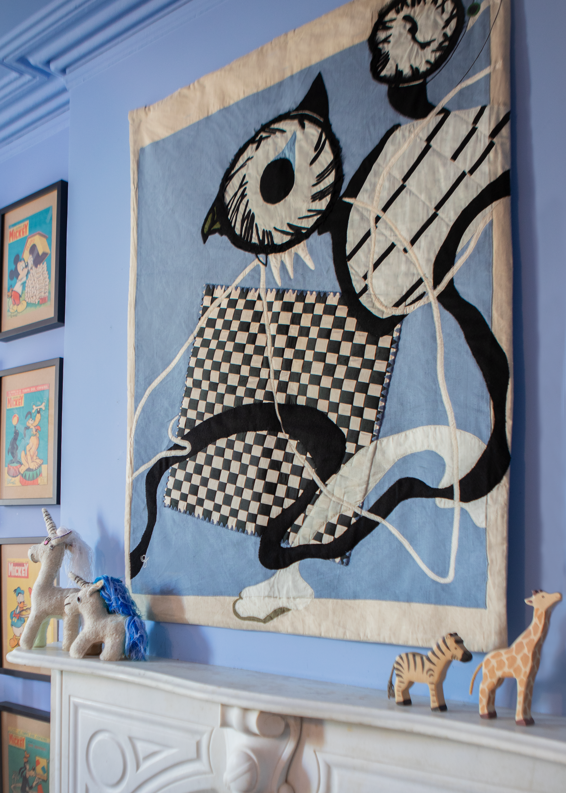 Hand made decorative tapestry with an abstract and surrealist design motif. Features a blue background with a checkerboard design and an abstract eye design. Hand crafted and hand embroidered from antique silks. Made in India, artisanal home decor. Modern tapestry design. Shown in a children's bedroom, styled with other modernist home decor and furniture. 