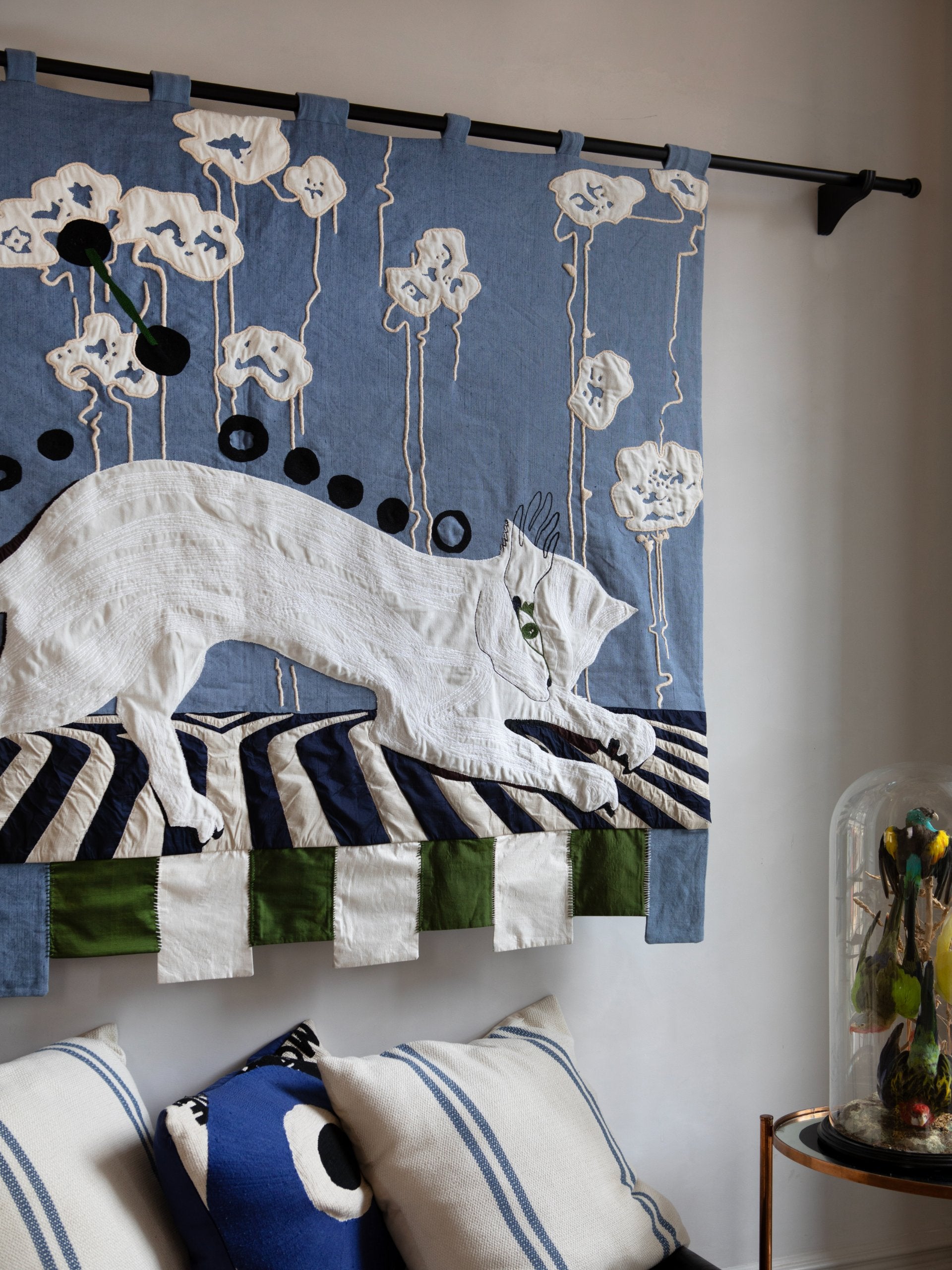 Detail of hand made decorative tapestry with an abstract and surrealist design motif. Features a cat design on a blue background with white flowers and a striped floor. Hand crafted and hand embroidered from antique silks. Made in India, artisanal home decor. Modern patchwork and applique tapestry design. Shown on a wall in a modernist styled home with other abstract home decor. 