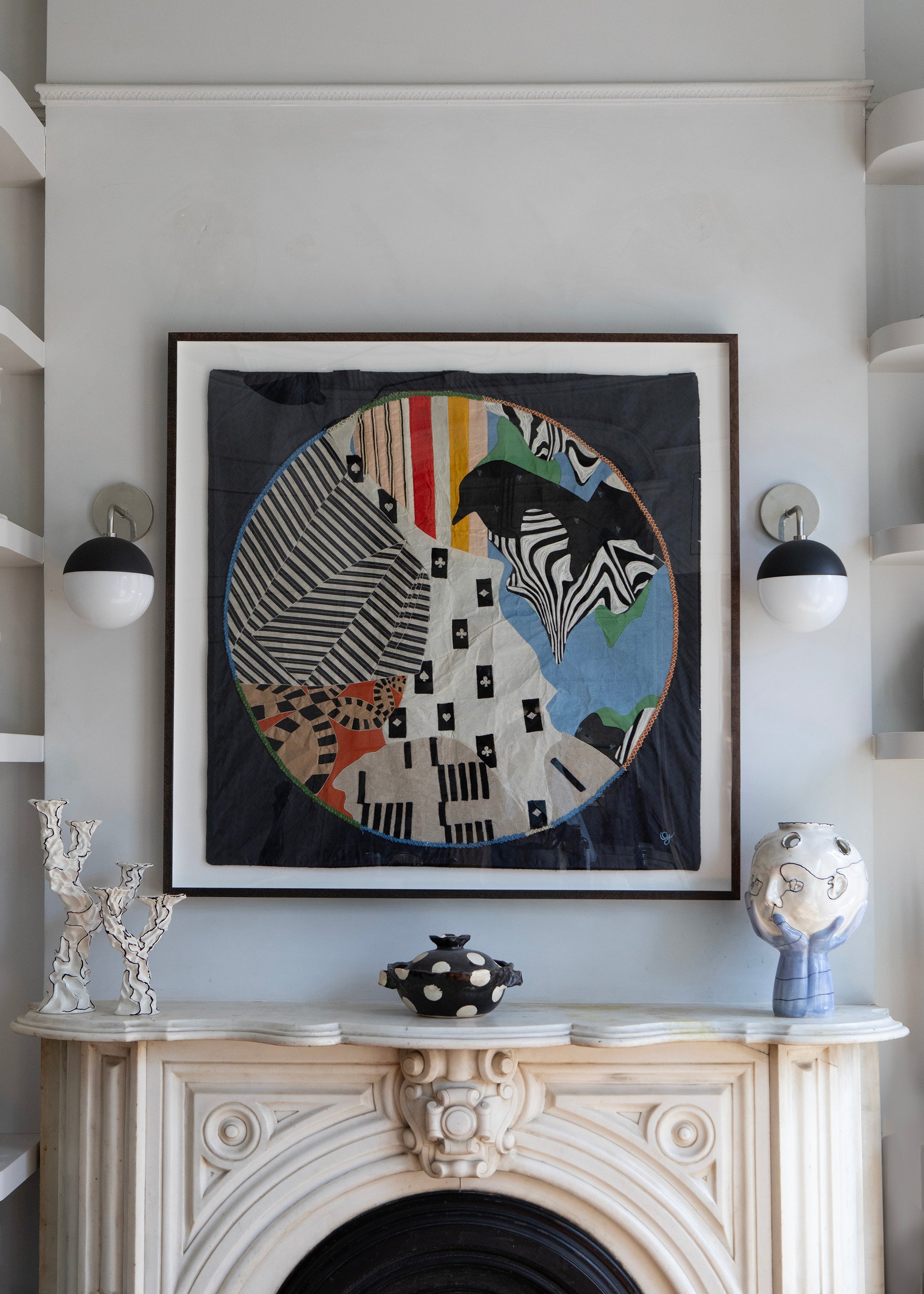Framed abstract artwork on a wall above a fireplace with decorative items. Hand made decorative tapestry with an abstract and surrealist design motif that suggests a globe. Hand crafted and hand embroidered from antique silks. Made in India, artisanal home decor. Modern tapestry design. Styled in a home setting with other modernist home decor and furniture. 