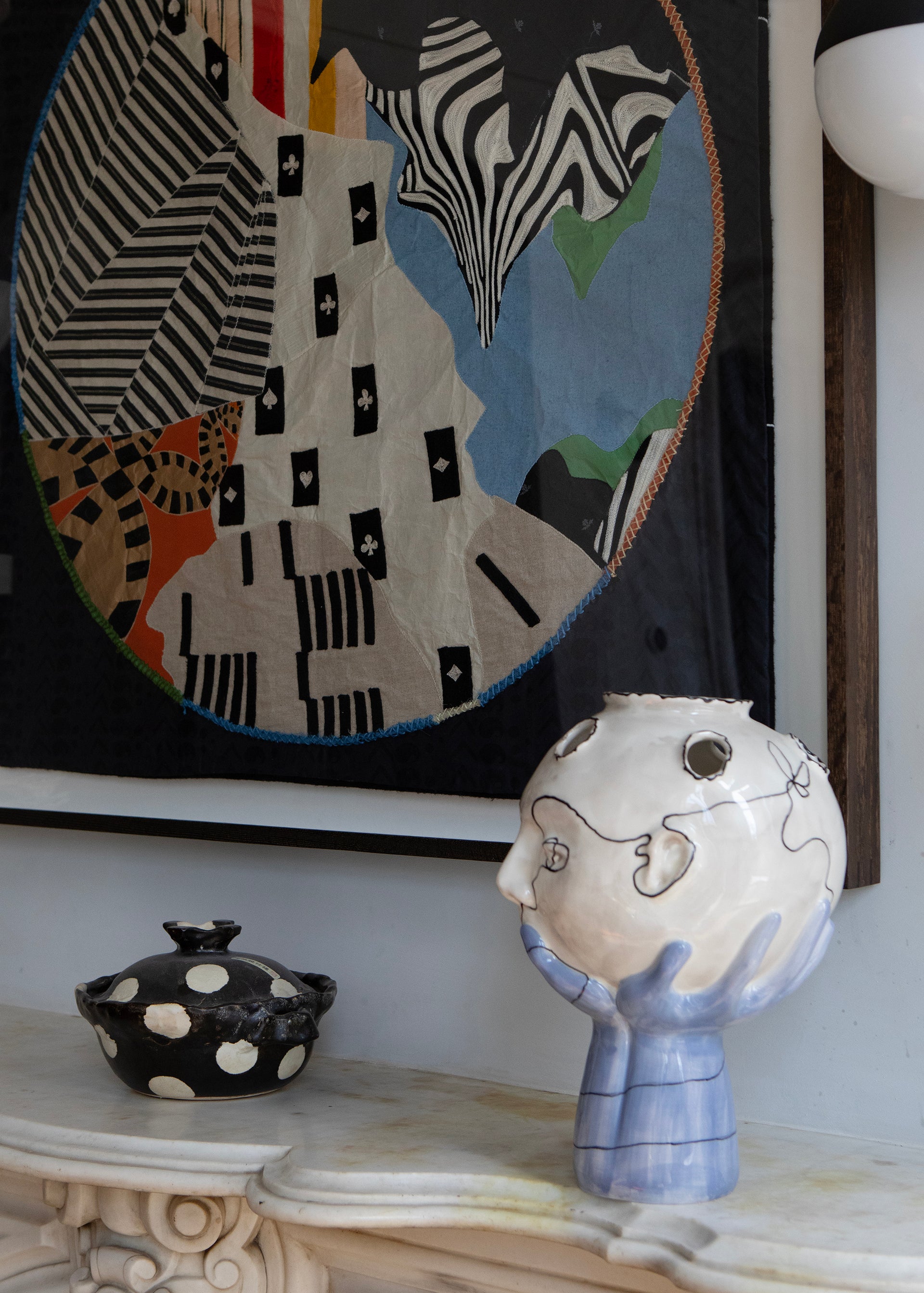 Framed abstract artwork on a wall above a fireplace with decorative items. Hand made decorative tapestry with an abstract and surrealist design motif that suggests a globe. Hand crafted and hand embroidered from antique silks. Made in India, artisanal home decor. Modern tapestry design. Styled in a home setting with other modernist home decor and furniture. 