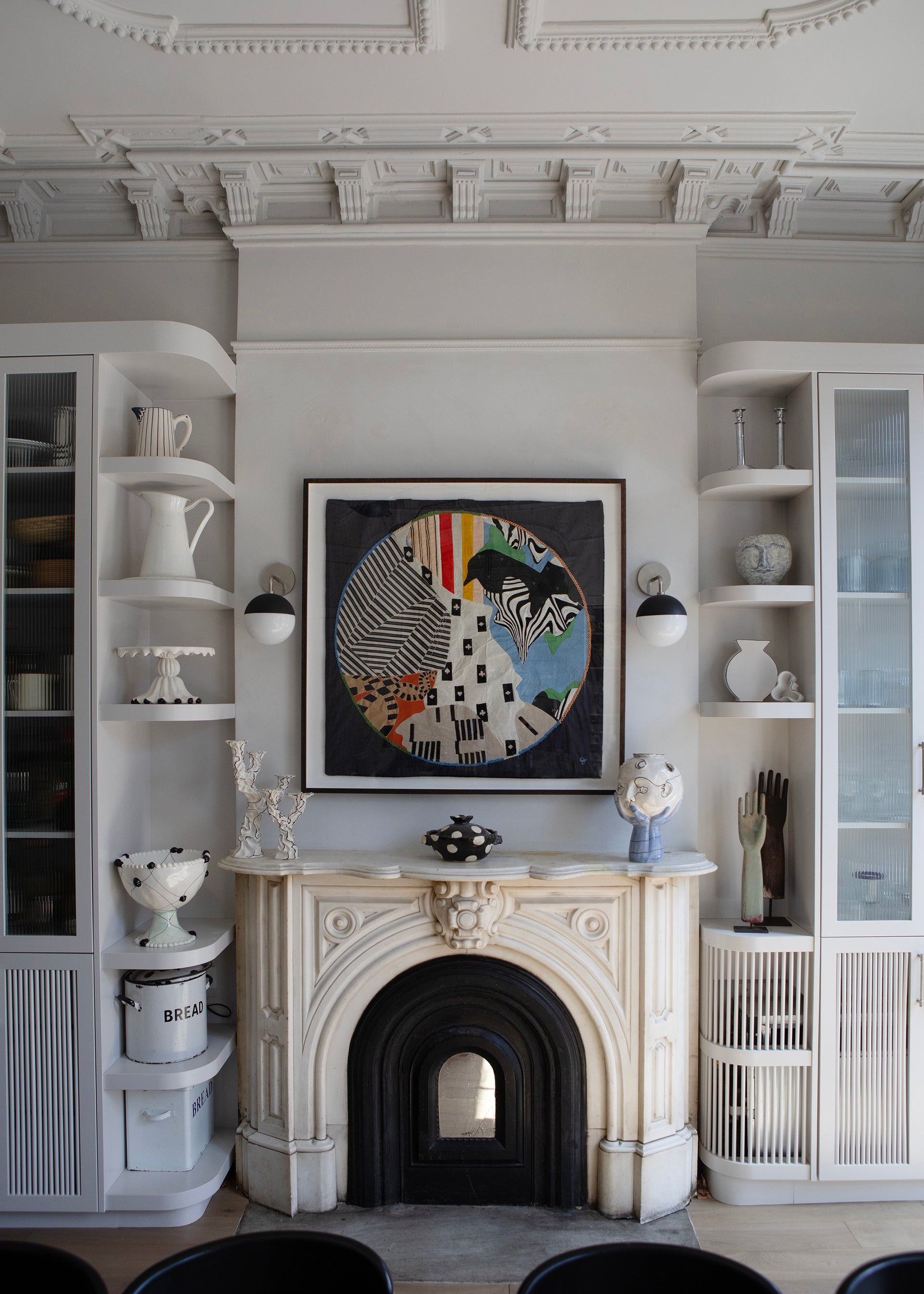 Framed abstract artwork on a wall above a fireplace with decorative items. Hand made decorative tapestry with an abstract and surrealist design motif that suggests a globe. Hand crafted and hand embroidered from antique silks. Made in India, artisanal home decor. Modern tapestry design. Styled in a home setting with other modernist home decor and furniture. 